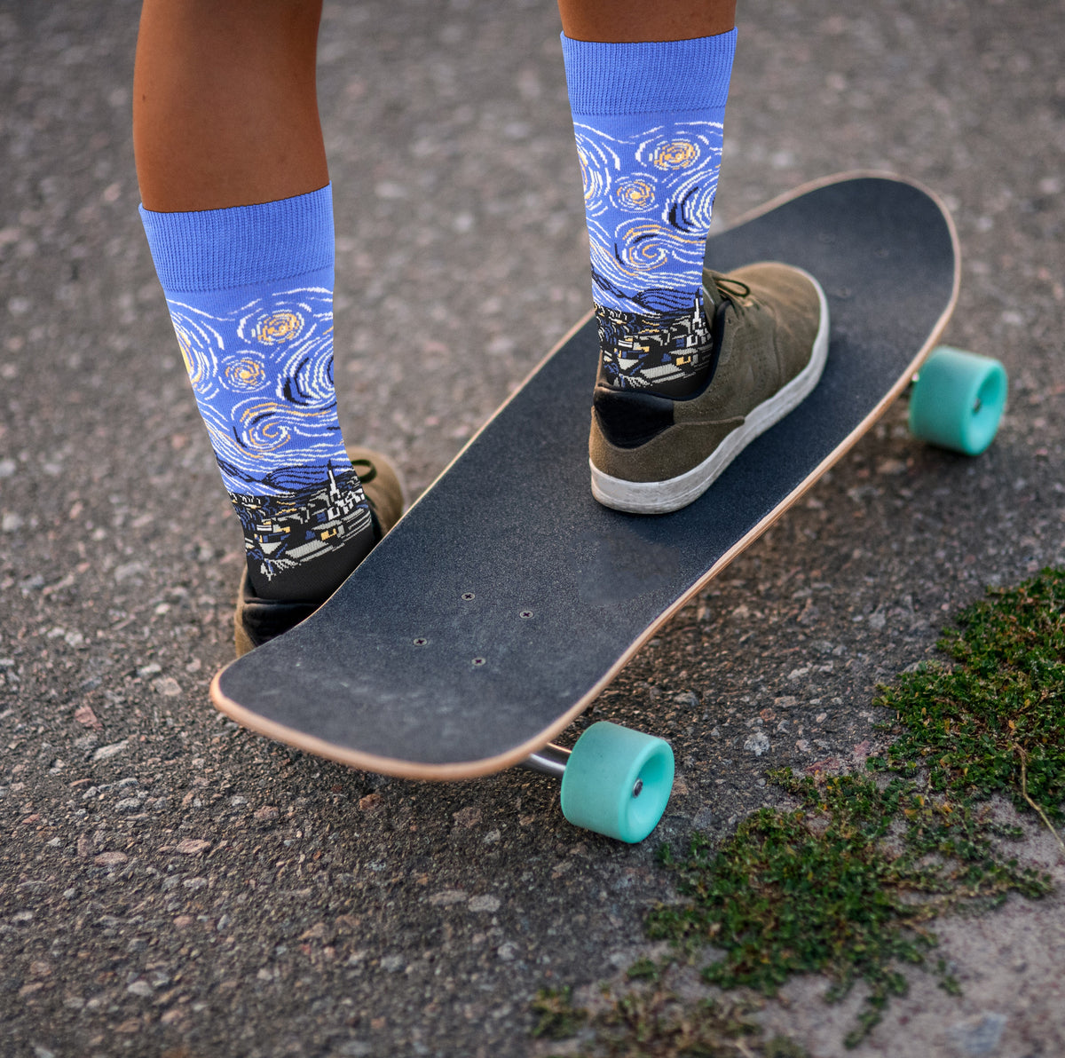 Starry Night socks worn with dark sneakers beside a skateboard with turquoise wheels.